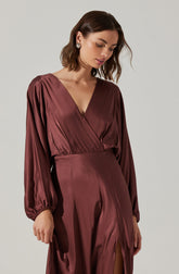 A woman is wearing a long, silky, deep burgundy dress with a V-neckline. The dress has long, puffed sleeves with elastic cuffs and a fitted waistband. There is a slit in the skirt showing some of her leg. She has short, wavy brown hair and is wearing statement earrings.